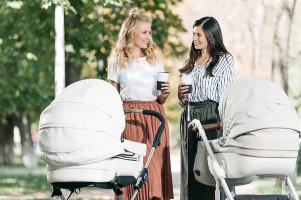 Mothers holding disposable coffee cups and looking at each other near baby strollers in park — Stock Photo