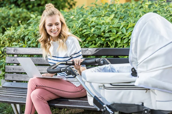 Pigiste travaillant avec ordinateur portable sur banc et tenant poussette bébé dans le parc — Photo de stock