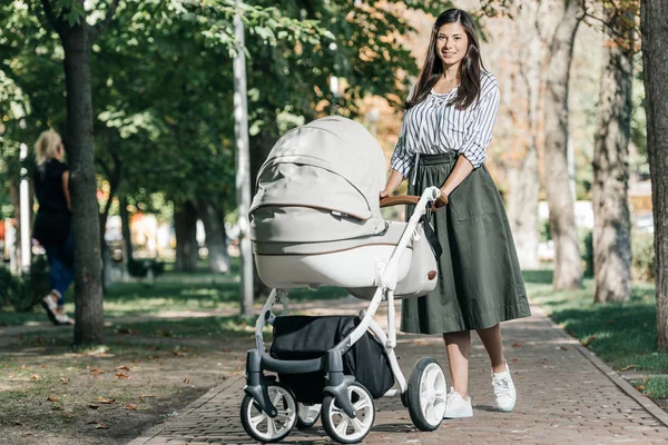 Beautiful mother walking with baby stroller in park and looking at camera — Stock Photo