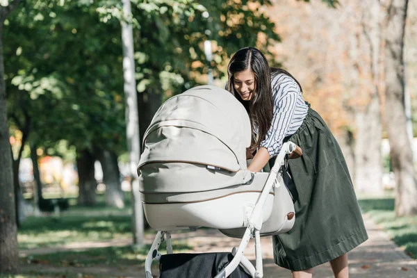 Attractive happy mother looking in baby stroller in park — Stock Photo