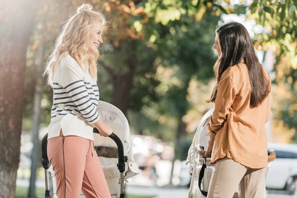 Side view of smiling mothers looking at each other while walking with baby strollers in park — Stock Photo