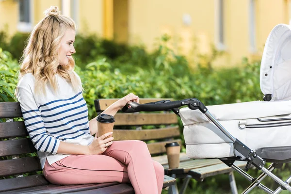 Vista lateral de mãe sorridente sentado no banco com café para ir e segurando carrinho de bebê no parque — Fotografia de Stock