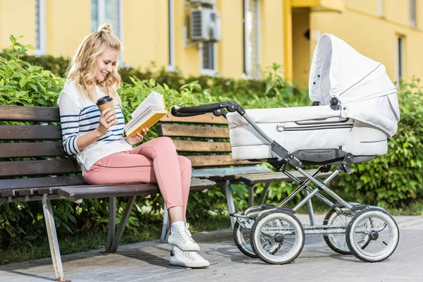 Smiling mother reading book and holding coffee to go on bench near baby stroller in park — Stock Photo