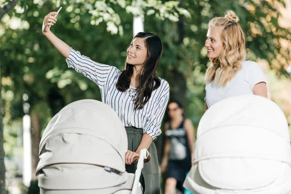 Happy mothers taking selfie with smartphone near baby strollers in park — Stock Photo