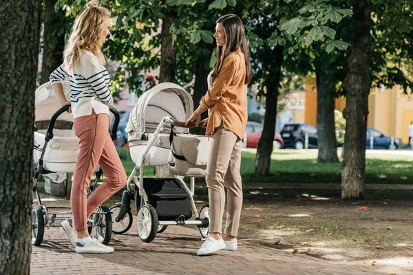 Visão lateral das mães conversando e de pé com carrinhos de bebê no parque — Fotografia de Stock