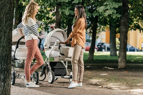 Vista laterale delle mamme che parlano e stanno in piedi con passeggini nel parco — Foto stock