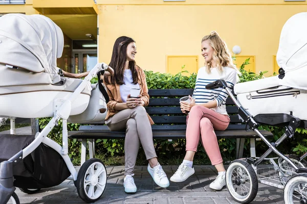 Mães sorridentes sentadas no banco com café para ir perto de carrinhos de bebê — Fotografia de Stock