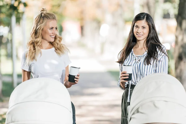 Mères souriantes debout avec poussettes bébé et café dans des tasses en papier dans le parc — Photo de stock