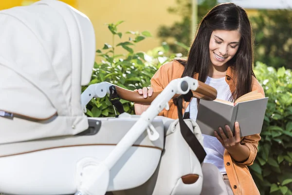 Sonriente madre lectura libro en banco cerca bebé cochecito en parque — Stock Photo