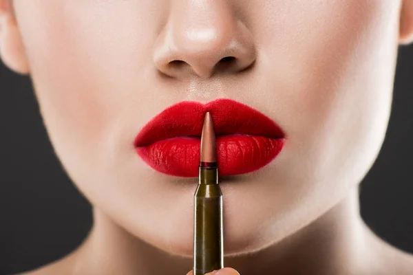 Vista parcial de una joven mujer sosteniendo una bala cerca de los labios rojos, aislada en gris - foto de stock