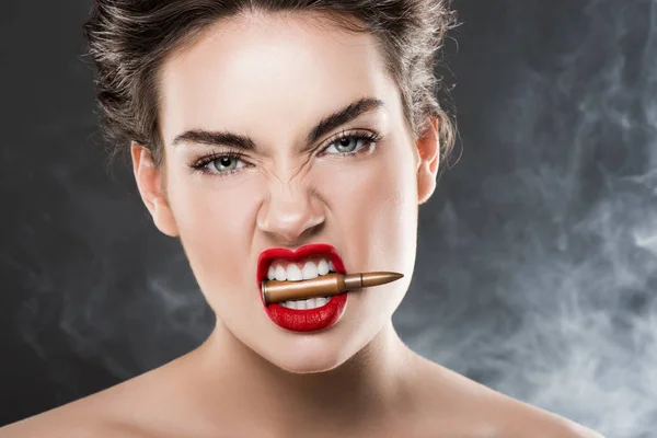 Mujer agresiva sosteniendo bala en los dientes, en gris con humo - foto de stock