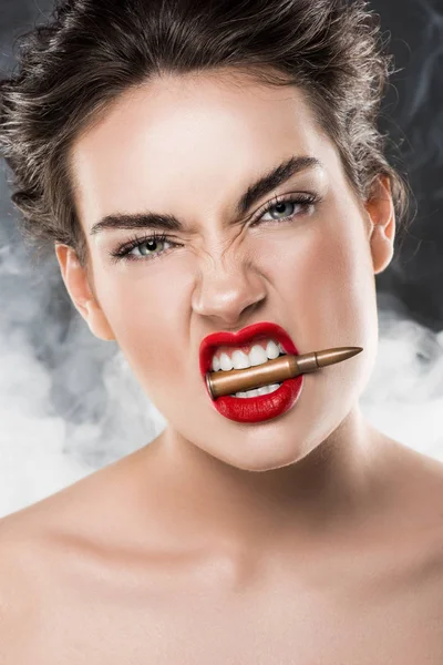 Bela mulher segurando bala nos dentes, em cinza com fumaça — Fotografia de Stock