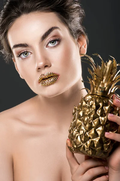 Beautiful girl with gild lips holding golden pineapple, isolated on grey — Stock Photo