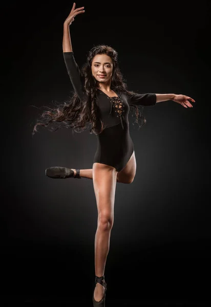 Bailarina sorridente em bodysuit preto e sapatos de balé dançando em pano de fundo escuro — Fotografia de Stock