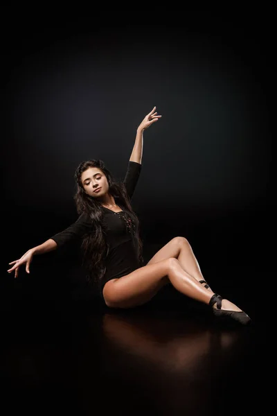 Joven bailarina elegante en traje de cuerpo negro y zapatos de ballet sentado en el suelo sobre fondo negro - foto de stock