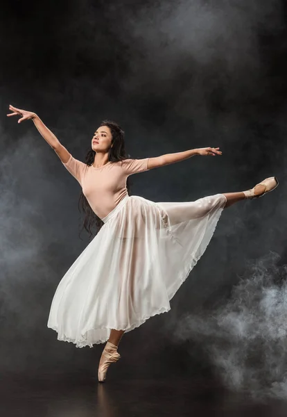 Bela jovem bailarina em saia branca dançando no fundo escuro com fumaça ao redor — Fotografia de Stock