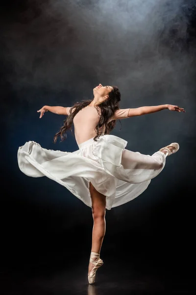 Beautiful young ballerina in white skirt dancing on dark background with smoke around — Stock Photo