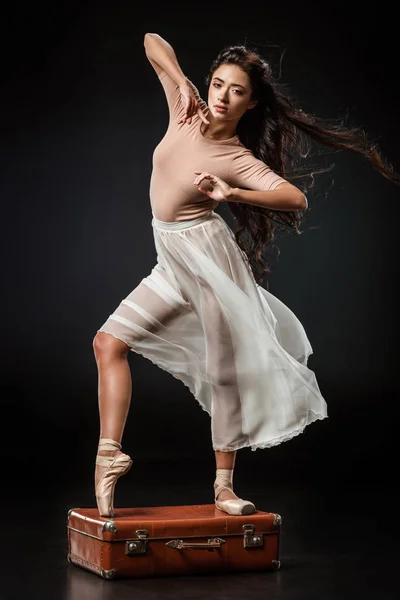 Молода елегантна балерина в білій спідниці позує на ретро валізі на темному фоні — стокове фото