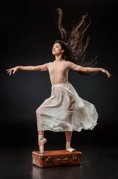 Young elegant ballerina in white skirt posing on retro suitcase on dark background — Stock Photo