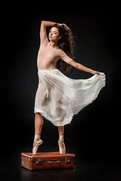 Young elegant ballerina in white skirt posing on retro suitcase on dark background — Stock Photo