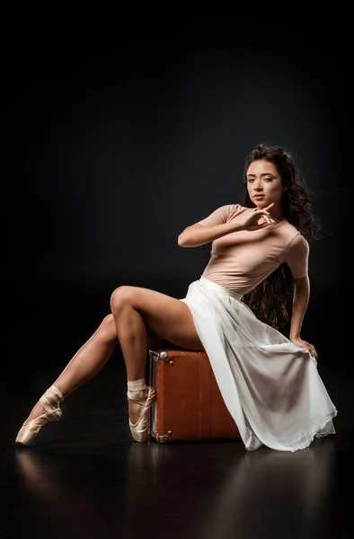 Side view of attractive ballerina in white skirt on retro suitcase on dark background — Stock Photo