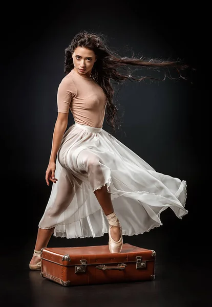 Bella ballerina in gonna bianca in piedi con una gamba su valigia retrò su sfondo scuro — Foto stock