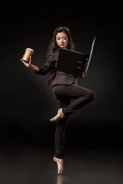 Atractiva mujer de negocios en traje y zapatos de ballet con carpeta y café para ir posando sobre fondo negro - foto de stock