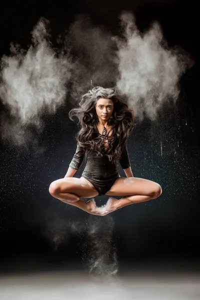 Belle ballerine en body noir sautant sur fond sombre avec talc en poudre autour — Photo de stock