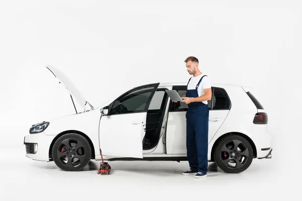 Schöner Automechaniker in blauer Uniform mit Laptop neben kaputtem Auto auf weiß — Stockfoto