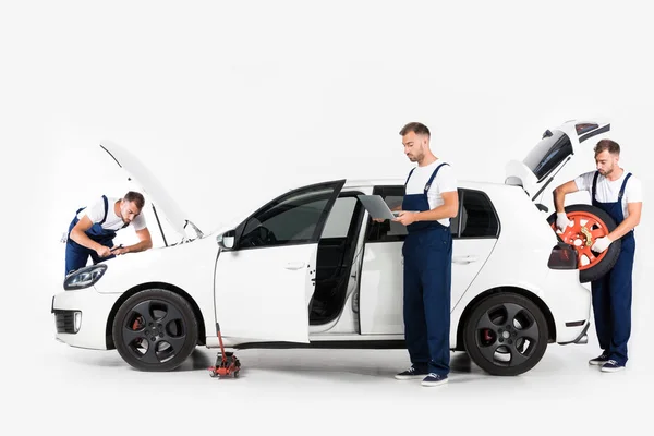 Mecánico automático teniendo neumático del coche, usando el ordenador portátil y mirando en la capucha abierta del coche en blanco - foto de stock