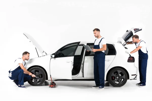 Auto mechanic changing car tire, using laptop and looking in open car trunk on white — Stock Photo