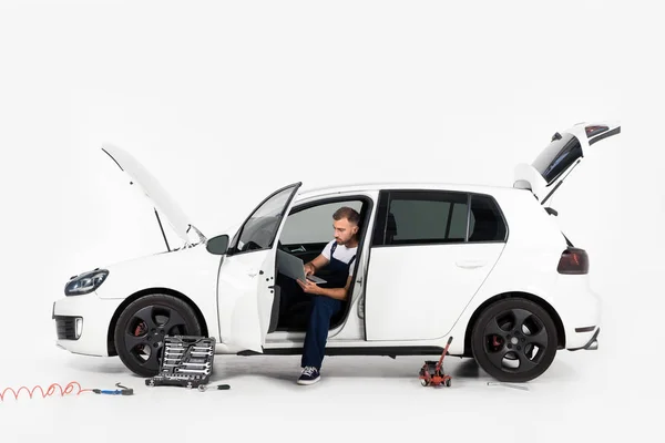 Mecánico auto guapo sentado en coche roto y el uso de la computadora portátil en blanco - foto de stock