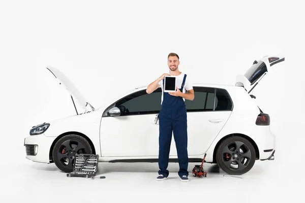 Lächelnder Automechaniker zeigt Tablet mit leerem Bildschirm neben kaputtem Auto auf weißem Grund — Stockfoto