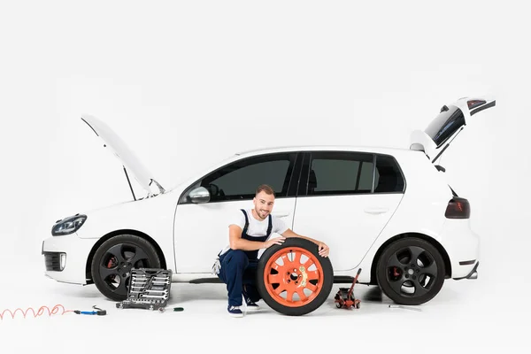 Smiling auto mechanic changing car tire and looking at camera on white — Stock Photo