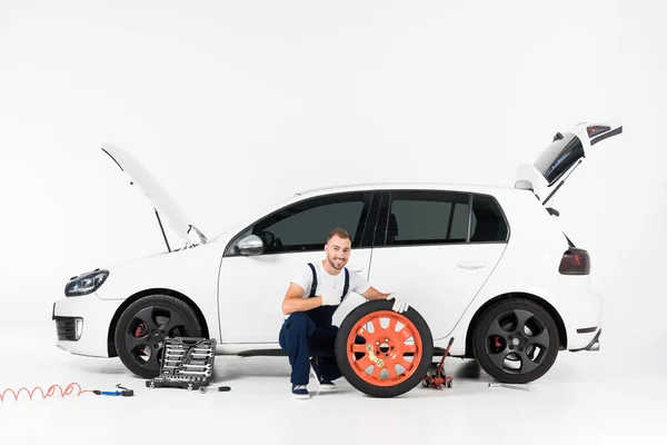 Auto mecânico agachamento perto do pneu do carro e mostrando polegar para cima no branco — Fotografia de Stock