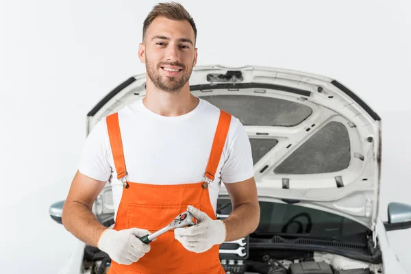 Smiling handsome auto mechanic holding monkey wrench near car on white — Stock Photo