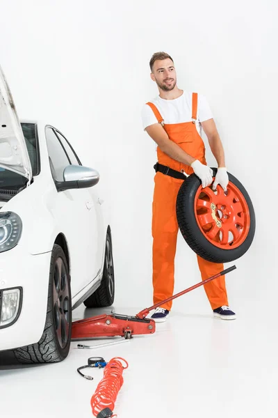 Meccanico auto in uniforme arancione che trasporta pneumatico auto su bianco — Foto stock