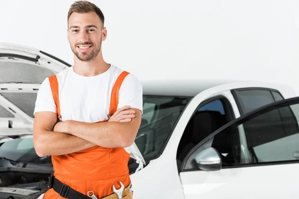 Mecánico auto guapo en uniforme naranja de pie con brazos cruzados cerca de coche abierto aislado en blanco - foto de stock