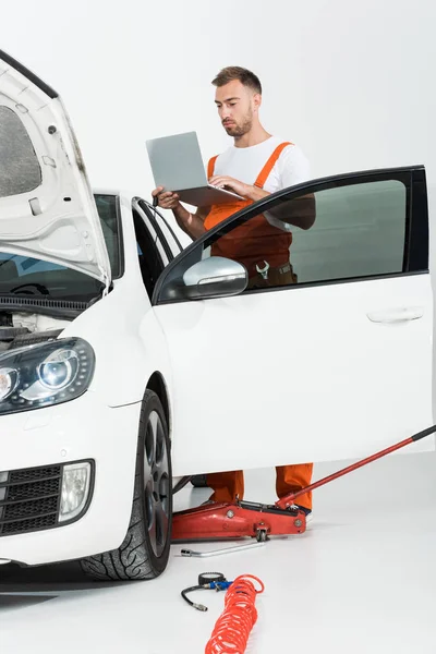 Bello meccanico auto in uniforme arancione in possesso di laptop vicino rotto auto isolato su bianco — Foto stock