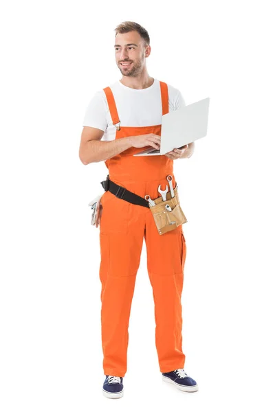 Mecánico auto guapo en uniforme naranja que sostiene el ordenador portátil y mirando hacia otro lado aislado en blanco - foto de stock