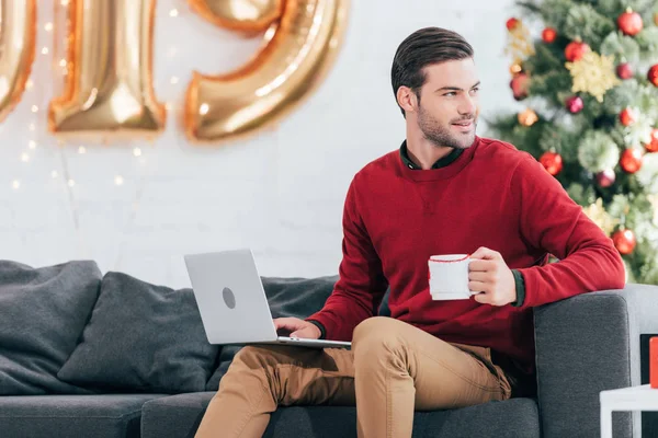 Hombre sonriente con taza de café usando el ordenador portátil en la víspera de Navidad — Stock Photo