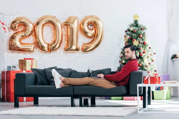 Homme utilisant un ordinateur portable sur le canapé à la maison avec arbre de Noël et 2019 ballons dorés — Photo de stock