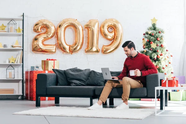 Homme avec tasse de café en utilisant un ordinateur portable à la maison avec 2019 ballons dorés et arbre de Noël derrière — Photo de stock