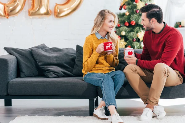 Bella coppia che tiene tazze di tè e si guarda a casa con albero di Natale — Foto stock