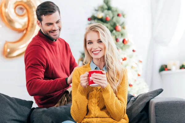 Beautiful happy woman holding coffee cup while man looking at her at home with christmas tree — Stock Photo