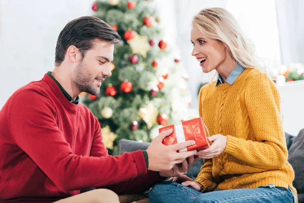 Man gifting present surprised to girlfriend at home with christmas tree — Stock Photo
