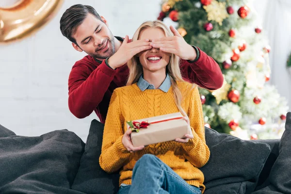 Junger Mann schließt die Augen und überrascht mit Geschenkbox für lächelnde Frau zu Hause mit Weihnachtsbaum — Stockfoto