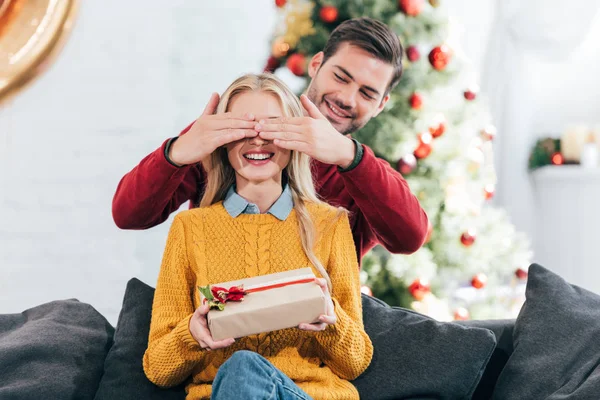 Hombre sonriente haciendo sorpresa con caja de regalo para mujer en casa con árbol de Navidad - foto de stock