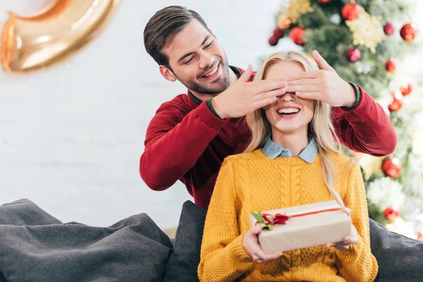 Schöner Mann schließt die Augen und überrascht mit Geschenkschachtel für Frau zu Hause mit Weihnachtsbaum — Stockfoto