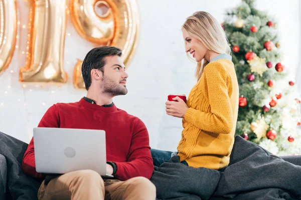 Mann mit Laptop schaut Frau mit Tasse Kaffee an, die mit goldenen Luftballons und Weihnachtsbaum auf dem heimischen Sofa sitzt — Stockfoto